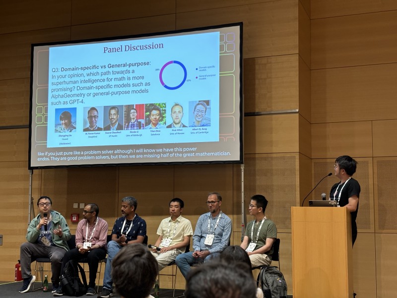 Six panelists sit on stage, in front of a slide titled &ldquo;panel discussion&rdquo;. A moderator stands behind a lectern to their right.