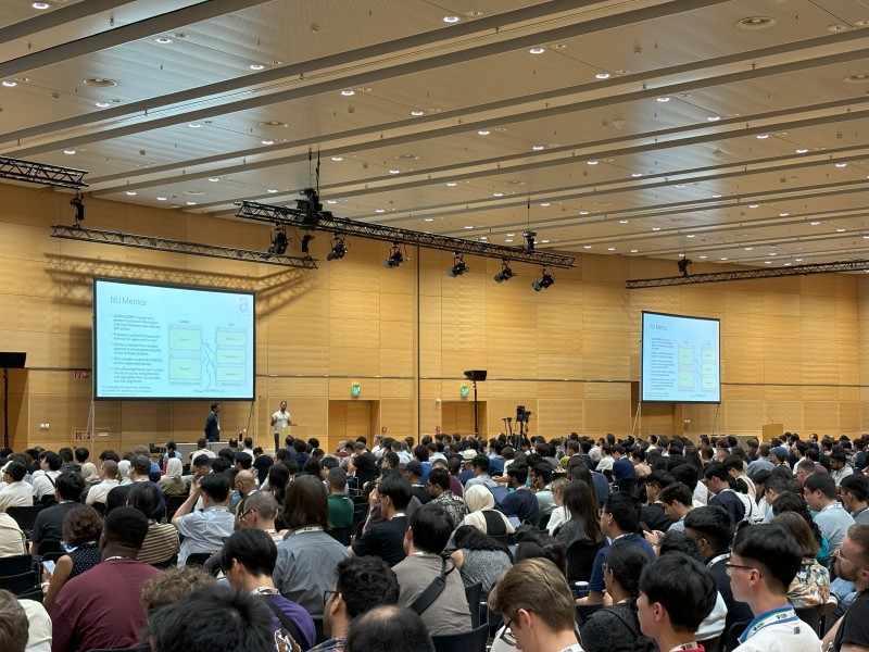 A full conference room with screens at the front showing a slide titled “NLI Metrics”