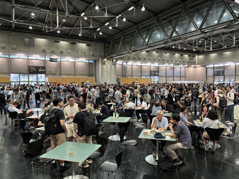 A packed hall with people sitting, standing, eating, and drinking