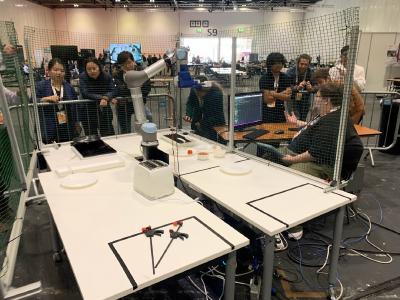 A robot arm set up on a table next to some plates, surrounded by people.